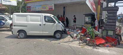 White van crashed into a storefront with scattered debris, including a red cart and bicycle; bystanders nearby.